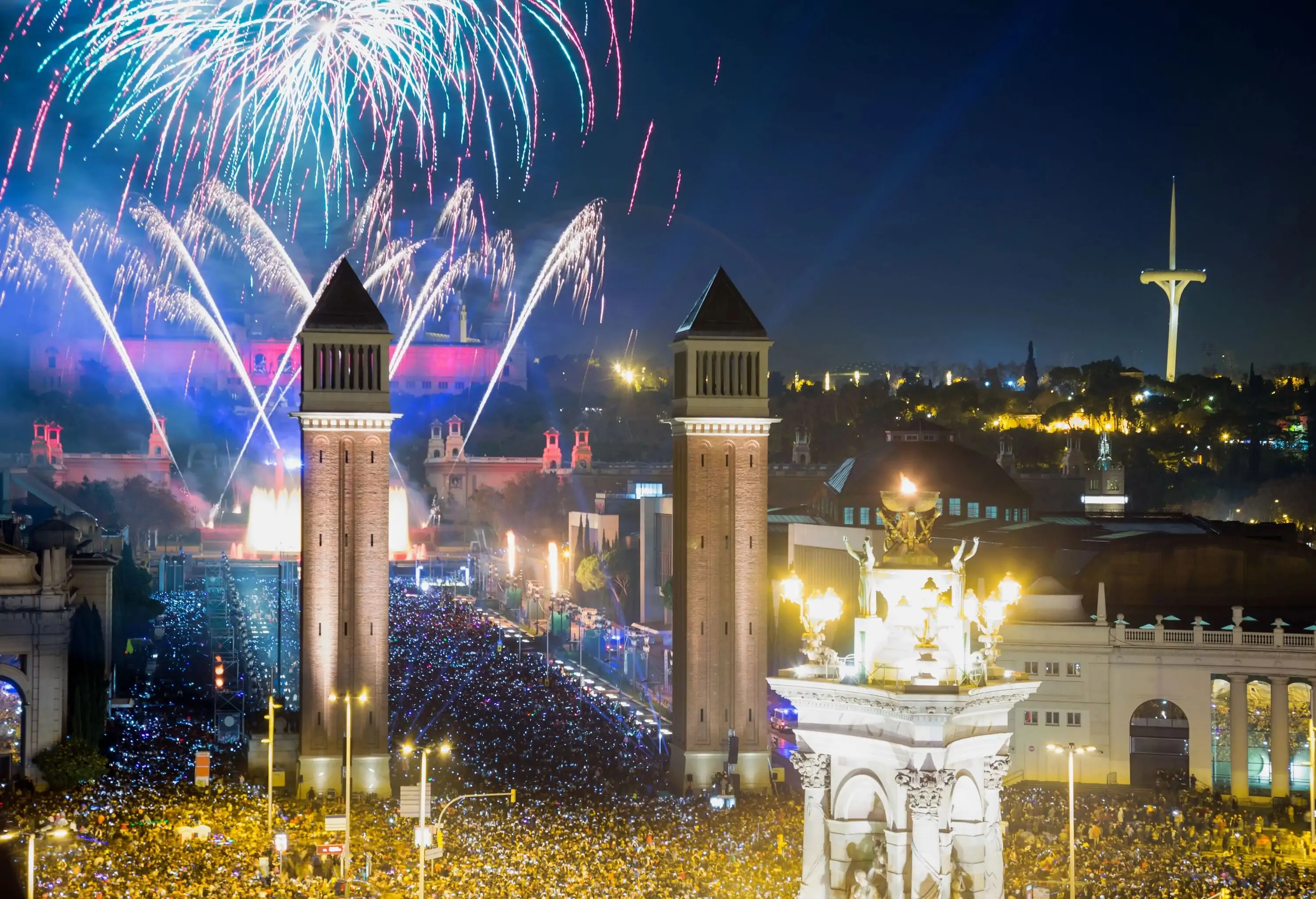 Skyline view of the crowded and brightly illuminated Plaça d'Espanya square with exploding fireworks in the night sky.